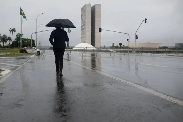 Imagem colorida de um homem caminhando em meio à chuva, usando guarda-chuva. Ao fundo, o Congresso Nacional