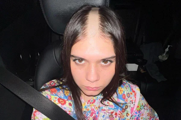 Mulher branca com cabelo louro e liso raspado no centro. Ela usa uma blusa estampada e colorida e está sentada em um banco de carro