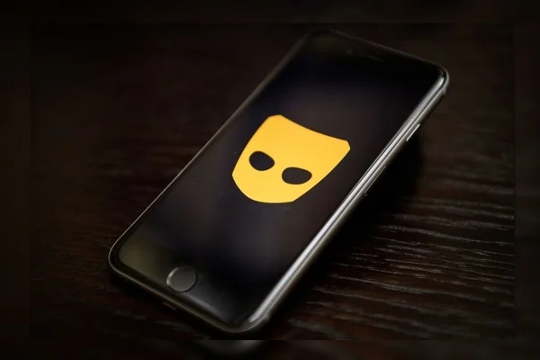 Imagem colorida de um celular, com o ícone do app Grindr no plano de fundo