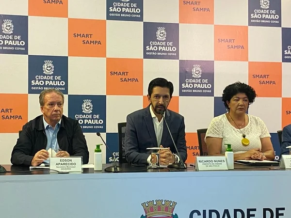 Imagem colorida mostra ricardo Nunes, homem branco de cabelo e barba pretos, de terno cinza e camisa branca, em uma mesa de auditório, ao lado de um homem branco, grisalho, e uma mulher negra de blusa branca, com um painel com logotipo da prefeitura ao fundo - Metrópoles