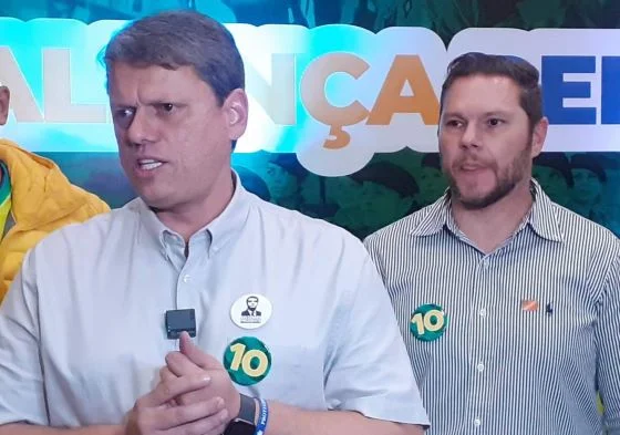 Imagem colorida mostra Tarcisio de Freitas e Danilo Campetti, homens brancos de camisa clara, em foto da cintura para cima na frente de um banner eleitoral - Metrópoles