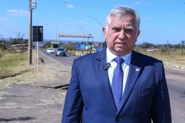 Imagem colorida do senador Izalci Lucas. Ele veste um terno azul, com gravata e camisa também azuis. No fundo, o Complexo Penitenciário da Papuda