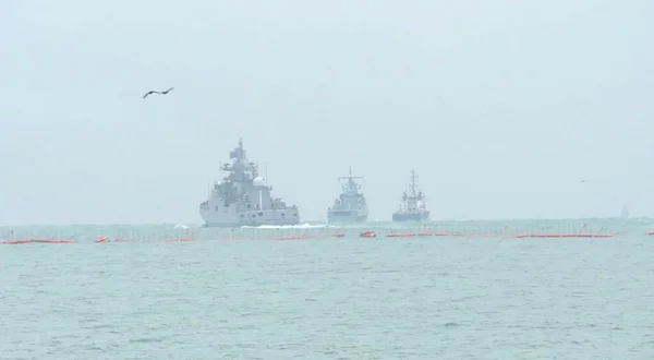 Um mar é mostrado sob muita névoa e céu turvo, com três navios militares atrás de uma contenção laranja. Eles estão mais distantes - Metrópoles