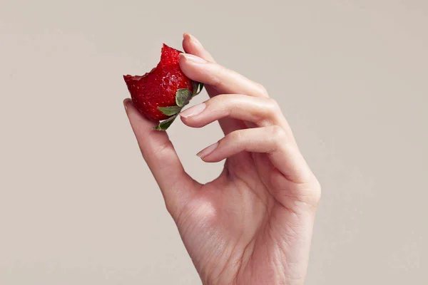Foto colorida de uma mão feminina segurando um morango mordido em um fundo bege - Metrópoles
