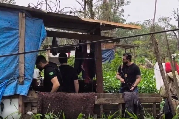 Foto colorida de movimentação de policiais em frente à barraco de madeira palco de homicídio.