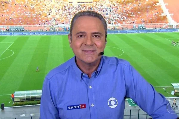 Foto de um homem branco em uma cabine de narração. Ele tem cabelo preto com grisalhos, usa blusa de longa azul e ao fundo um campo de futebol - Metrópoles