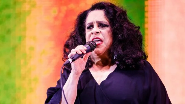Foto colorida de Gal Costa. ela está cantando com o microfone na mão e utiliza uma roupa preta. Ele também tem o cabelo preto - Metrópoles