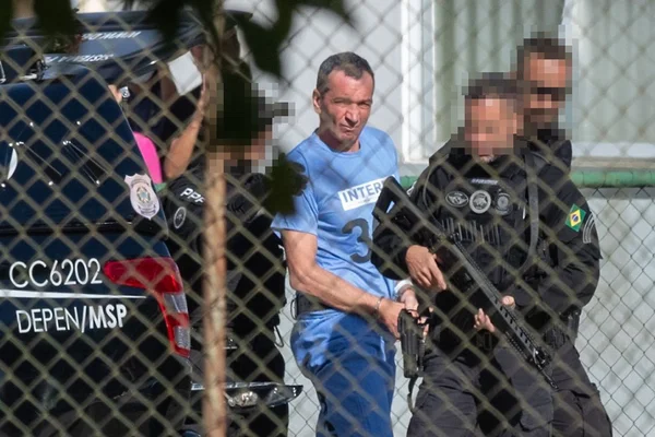 Marcola, homem branco, magro, de cabelos curtos e escuros, veste uniforme azul, usado na prisão; ele é escoltado por policiais de uniformes pretos e com rostos borrados; os policiais caminham ao lado de Marcola, e o grupo aparece próximo a um carro da polícia -- Metrópoles