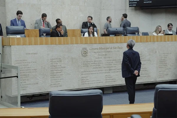 Imagem mostra o plenário da Câmara Municipal de São Paulo, com vereadores sentados e um homem em pé voltado à mesa. Ambiente interno, iluminação clara e neutra, painel de mármore bege com nomes gravados - Metrópoles