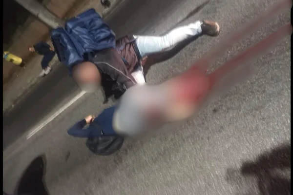 Imagem colorida, com desfoque da cintura para baixo de mulher caída em asfalto sendo amparada por motoqueiro com mochila de entrega azul - Metrópoles