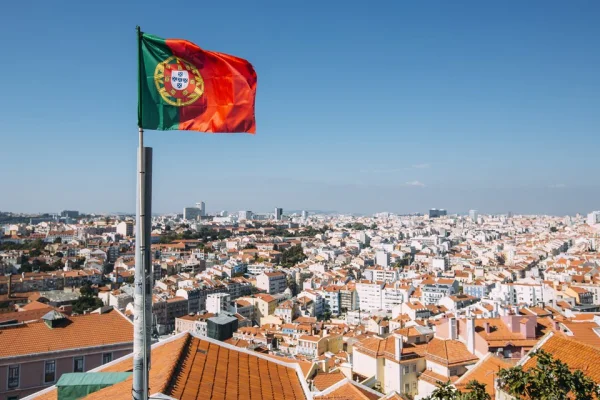 Foto colorida da bandeira de Portugal e há construções em volta do mastro