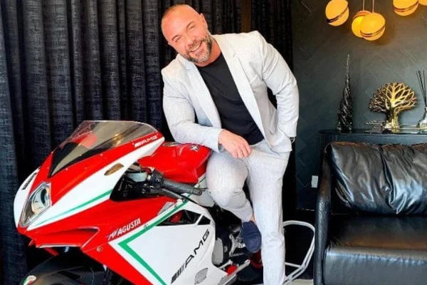 Homem apoiado em moto dentro de casa, sorrindo - Metrópoles