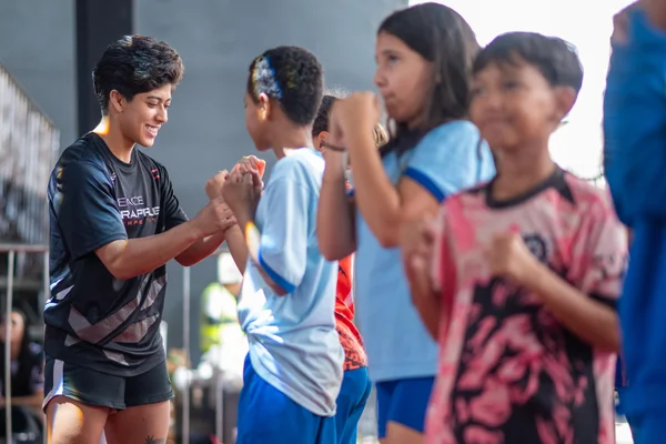 Três crianças ficam lado a lado segurando luvas de treino. Elas interagem umas com as outras, sorrindo e gesticulando. O ambiente é amplo, com fundo luminoso e estrutura metálica.