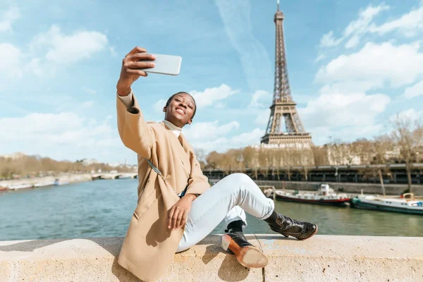Mulher negra está sentada na ponte sobre o rio Sena com a Torre Eiffel, em Paris, ao fundo tirando uma selfie