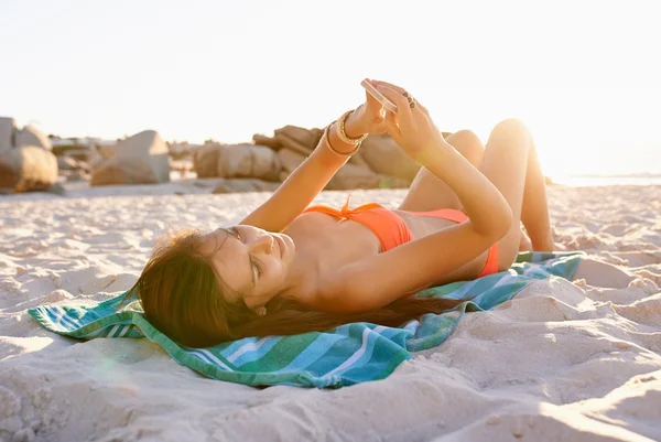 Foto de uma jovem usando seu celular deitada em uma toalha de praia tomando sol. Metrópoles
