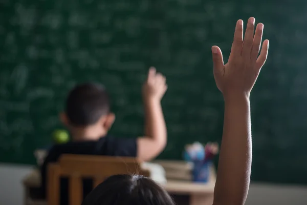 Foto colorida com zoom e falta de foco de mão infantil levantada em sala de aula - Metrópoles.