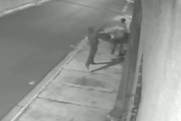 Imagem colorida de homem jogando pedra na cabeça de outro no meio da rua. Metrópoles