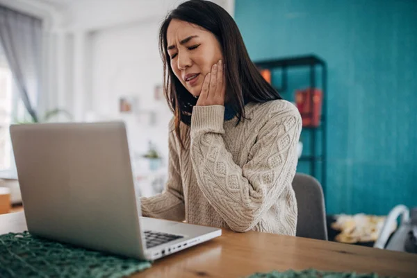 Foto de mulher sentada em frente a um computador com uma mão no rosto e cara de dor - Metrópoles
