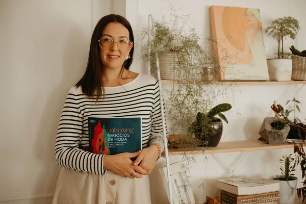 Marília Caravalhinha, uma mulher branca de cabelos escuros, usando blusa listrada e óculos de grau. Ela segura um livro colorido - Metrópoles