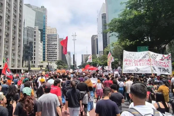 Imagem colorida mostra aglomeração de pessoas segurando faixas vermelhas em avenida - Metrópoles