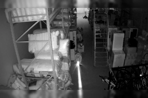 Imagem em preto e branco de um depósito de um estabelecimento comercial, com quatro homens engatinhando no chão antes de furtar o local. Na imagem, há mercadorias embaladas