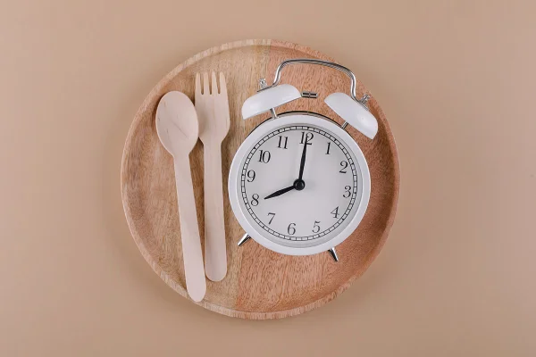 Despertador em prato de madeira com talheres. Conceito de jejum intermitente, dieta e perda de peso. Metrópoles