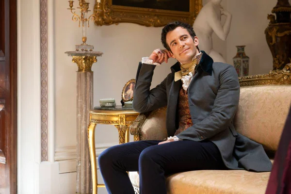 Na imagem colorida, homem está sentado em sofá e sorrindo, com roupa de estilo vitoriano - Metrópoles