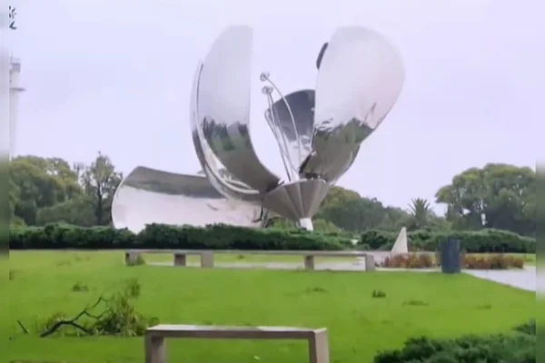 Foto colorida mostra paisagem de gramado com uma flor de aço metalizada no seu centro destruída pelos videos. Floralis Genérica
