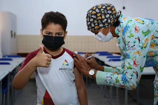 Aluno da rede básica de educação de Aparecida (GO) recebe segunda dose da vacina contra COVID-19 de enfermeria e faz sinal de joia com a mão - Metrópoles