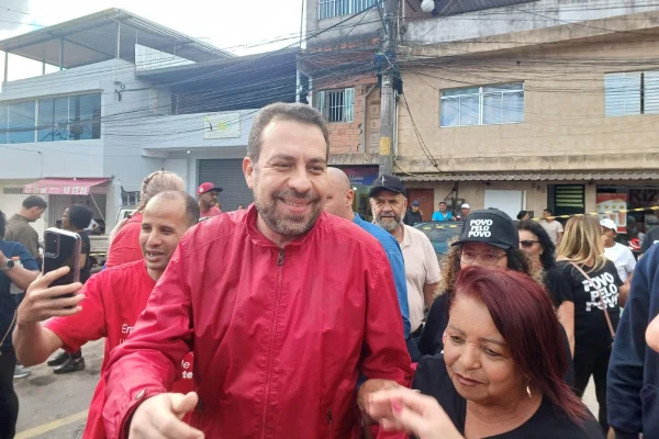 Imagem colorida mostra homem de cabelo curto escuro e barba também escura. Ele veste uma jaqueta fechada na cor vermelha e estica as duas mãos à frente do corpo. Ele está sorridente, é Guilherme Boulos -- Metrópoles