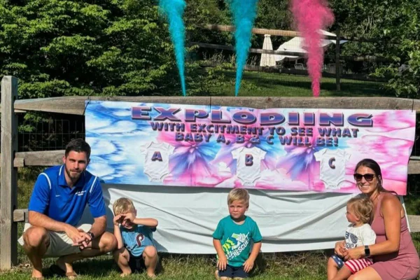 Casal posa com três filhos em frente a faixa e fumaça colorida de chá revelação