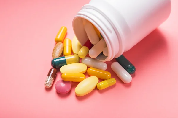 Foto mostra frasco branco de onde saem pílulas coloridas e cápsulas de multivitamínicos em um fundo rosa pastel. Exames vitaminas