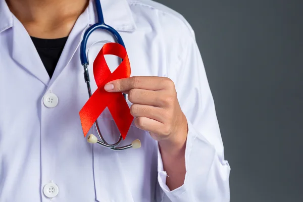 Foto colorida de pessoa vestida de branco com estetoscópio no pescoço, segurando fita vermelha que simboliza campanha nacional contra HIV - Metrópoles.