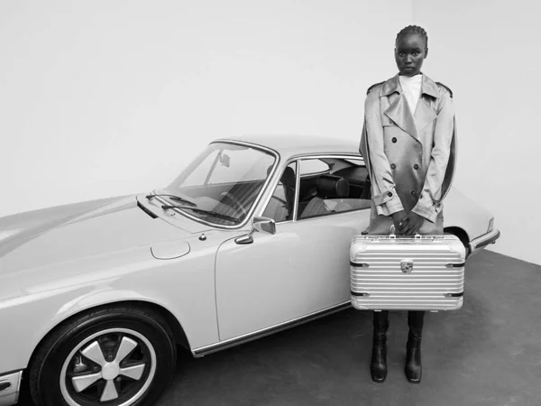 Na imagem em preto e branco, modelo posa em frente de carro conversível segurando mala da Rimowa com a Porsche