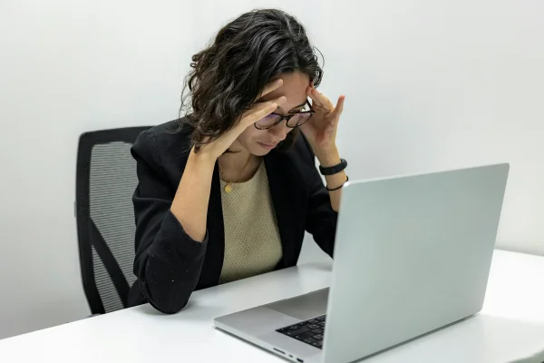 Foto colorida de mulher sentada e com notebook à frente em uma mesa. Ela está com as mãos no rosto - Metrópoles