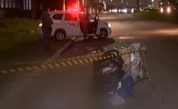 Imagem colorida, pouco nítida, de carrinho de recicláveis caído, isolado por faixa zebrada. Ao fundo, uma viatura da PM - Metrópoles