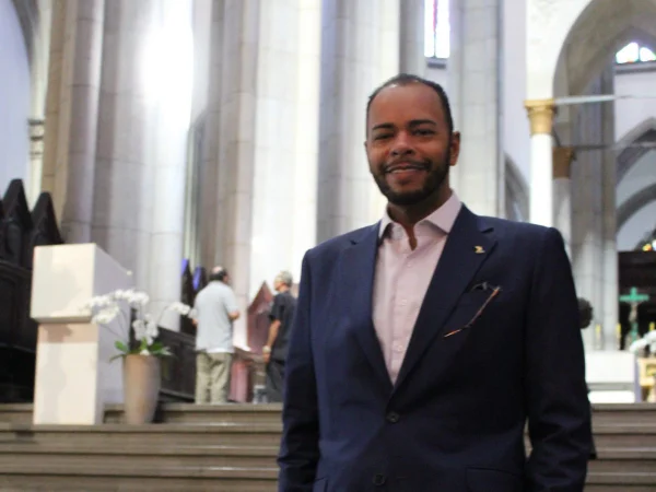 Imagem colorida mostra Fernando Alfredo, homem negro, de cabelos pretos e barba aparada, da cintura para cima, vestindo terno azul e camisa rosa, à frente da escadaria do altar da igreja da Sé - Metrópoles