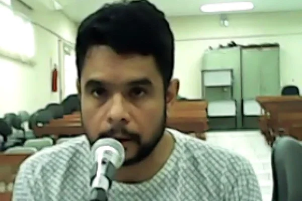 Imagem colorida de João Paulo Primus Fernandes da Costa durante audiência. Ele é um homem pardo, de barba, e fala no microfone do tribunal durante julgamento por ser falso médico - Metrópoles