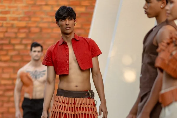 A imagem mostra um modelo desfilando. Vestindo um conjunto de camisa e calça vermelha, ele caminha com acessórios na cintura