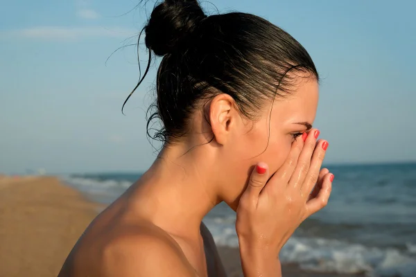 Imagem mostra perfil de mulher de cabelos presos, com as mãos no rosto. Ela está em uma praia - Metrópoles