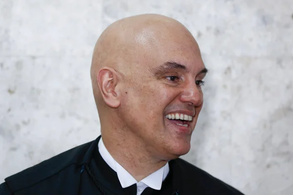 Imagem colorida mostra o rosto de Alexandre de Moraes, homem branco, careca, e toga preta, em um fundo cinza. Ele olha para sua esquerda e sorri - Metrópoles