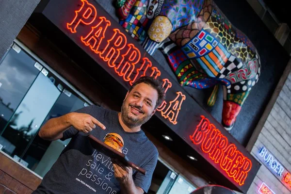 Chef Gil Guimarães em frente ao Parrilla Burger segurando um hambúrguer