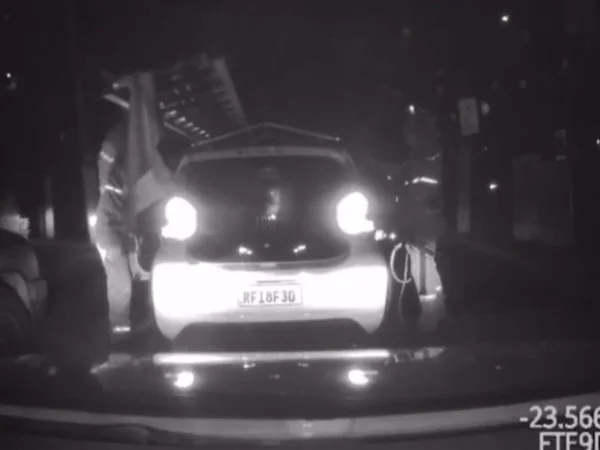 Imagem em preto e branco mostra um carro branco com uma escada em cima. Em cada um dos lados do veículo, há um homem com uniforme da empresa de energia elétrica.