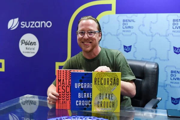 Foto colorida de um homem loiro chamado Blake Crouch segurando três livros coloridos - Metrópoles