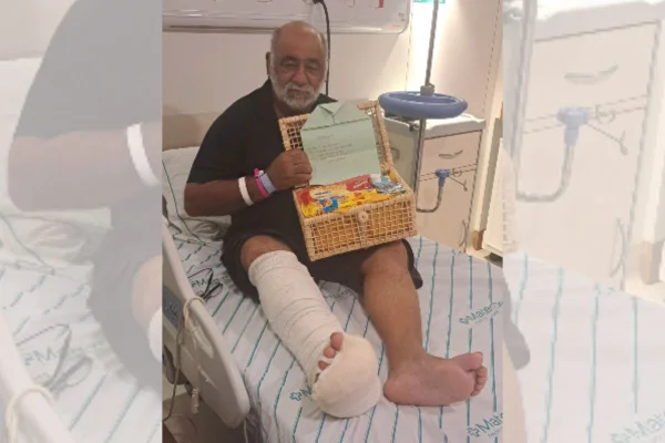 Imagem colorida mostra Nuremberg José Maria em uma cama de hospital com o pé enfaixado e segurando uma cesta de produtos | Metrópoles