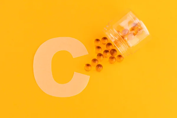 Foto colorida com fundo laranja, a imagem da letra C e um pote transparente com cápsulas redondas de vitamina C - Metrópoles.