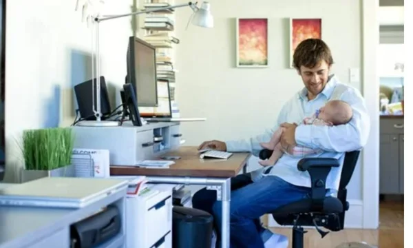 Imagem de homem, em sua casa, sentado na cadeira do escritório, segurando o filho, um bebê, enquanto trabalha. O homem veste uma camisa branca e calça jeans azul e olha para a criança enquanto mexe no mouse - Metrópoles