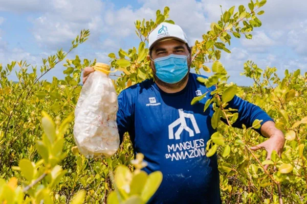 homem com boné e camisa azul segura uma garrafa de plástico em meio a plantas