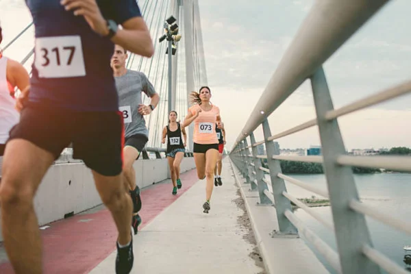 Foto em perspectiva mostra corredores em movimento sobre uma ponte, com guarda-corpo metálico à direita. O sol ilumina o percurso, destacando a silhueta dos atletas