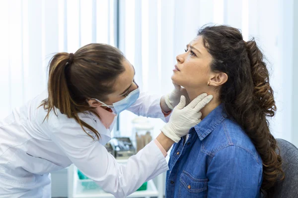 Imagem mostra uma mulher de jaleco, luvas e máscara analisando o pescoço de uma paciente, que está olhando para cima - Metrópoles